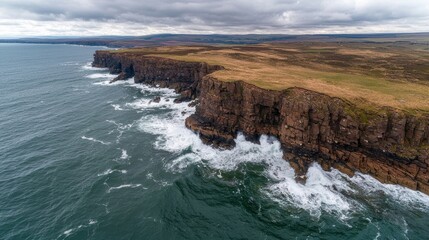 Aerial view of rugged cliffs meeting the ocean, showcasing dramatic rock formations and crashing waves against a serene, cloudy sky.