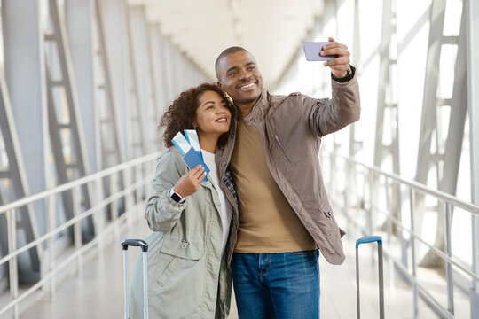 Travel Memories. Cheerful african american Couple Taking Selfie On Smartphone In Airport, Holding Tickets, Waiting For Boarding, Free Space