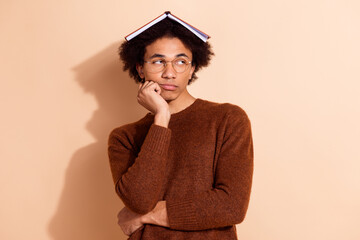 Young African American student with a book on his head looking thoughtful against beige background