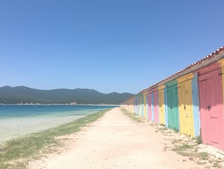 Colorful beach huts line a sandy path by the clear blue water.