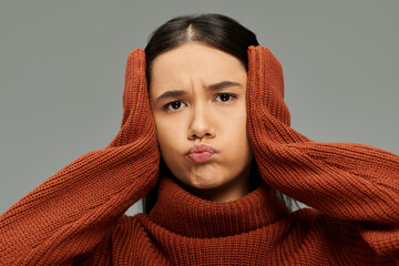 A young woman displays her emotions with a pout, hands on her head, in a cozy setting.