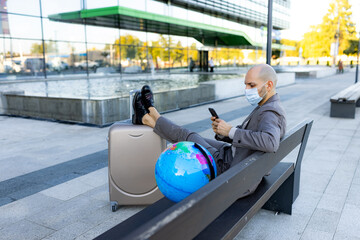 A man wearing a face mask sits on a bench with a globe and suitcase, representing travel readiness and caution in a contemporary urban environment.