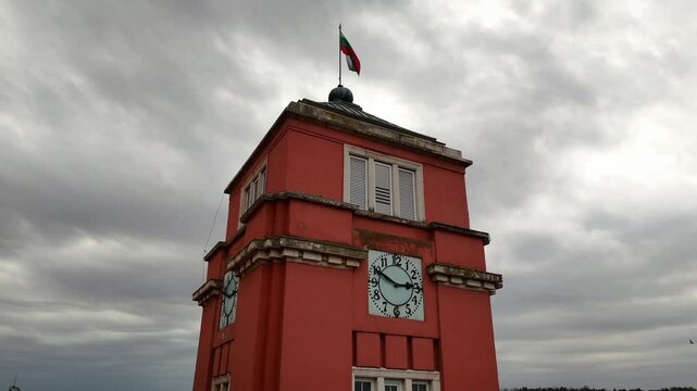 the clock tower with flag on its top and birds flying near, under a cloudy sky in Pleven, Bulgaria