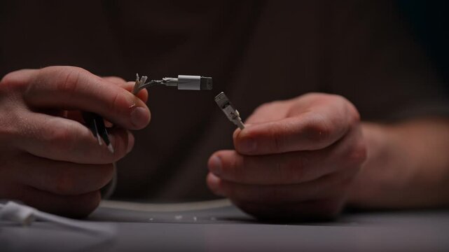 Man looking, inspecting, examining broken white USB lightning cable, showcasing intricate process of restoring damaged electronics, at table in workshop with dark interior, closeup, slow motion.