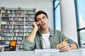 A dedicated student takes notes while on a call in a modern library filled with books.