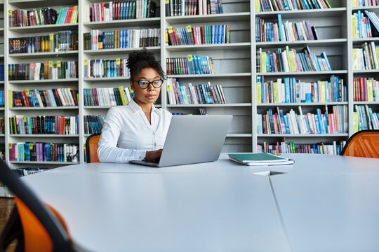 An inspiring teacher works on her laptop in a colorful library full of books and resources.