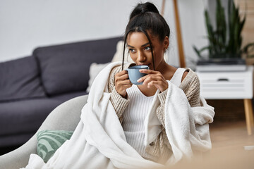 A young woman wrapped in a blanket enjoys a warm drink while focused on her studies at home.