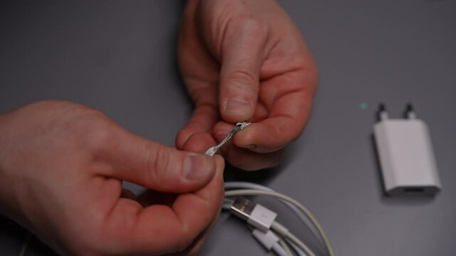 Top view of technician man carefully inspecting and examining damaged white USB cable, showcasing intricate wiring and delicate repair process for restoring connectivity in workshop with dark interior