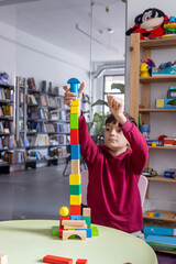 A young boy builds a tower with wooden blocks in a children’s room, highlighting themes of...