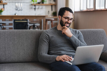 Arabian man is casual clothes sitting couch in a modern apartment smiling looking at his laptop....