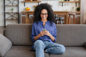 Smiling young African woman using cell phone relaxing on couch at home. Happy lady sitting on sofa...
