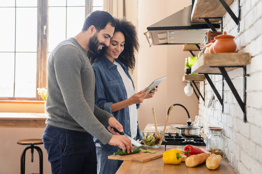 Joyful young mixed race couple african woman arabian man using online app on tablet cooking together in home kitchen ordering food delivery on supermarket store internet website consulting recipe - Powered by Adobe