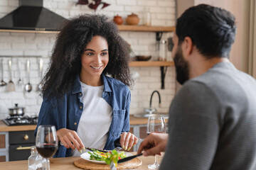 Laughing happy mixed race millennial african wife and arabian husband eating salad drinking red wine in light kitchen interior. Romantic dinner at home together. Love relationship family