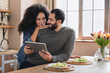 Young mixed race couple using digital tablet while have healthy dinner in light modern kitchen,...