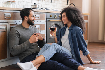 Close up of African Arabian couple drinking red wine sitting kitchen floor at home happy wife and husband satisfied customers celebrating relocation in new apartment mortgage