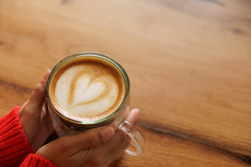Hands of a woman holding a cup of coffee. Latte art, heart drawn in coffee, caffeine for energy. Customer, student or businessman having breakfast in a store or cafe.
