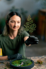 Close-up in a large green workshop, a woman with green hair in black gloves holds a sprig of green...