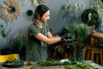 in large green workshop a thin woman with green hair in black gloves at a large table collects a...