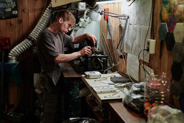 Young repairman in casual attire and eyeglasses using handtools while fixing details of industrial machine in workshop