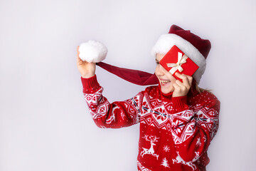 Cheerful child in a festive sweater holding a gift while wearing a Santa hat against a plain...