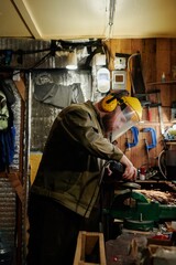 Young bearded metalworker in protective mask and headphones using electric saw while grinding metal detail