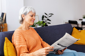 Smiling senior woman wrapped in a warm blanket reading newspaper on sofa at home in winter season