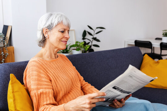 Smiling senior woman wrapped in a warm blanket reading newspaper on sofa at home in winter season