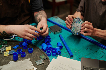 Hands of male technicians of recycling factory sitting by workplace with plastic supplies and...