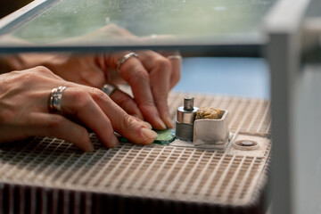 close up in a large green workshop, a thin woman with green hair at a wooden table on a grinding machine palates the ends of a glass product, polishing the corners, handmade