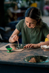 in a large green workshop, a woman with green hair creates decorative flowers laying them out on a round mirror, handmade with a soldering iron.