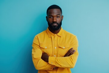Obraz premium Young African American man in his 20s wearing a yellow shirt arms crossed posing for a studio portrait against a light blue background Lifestyle theme