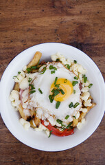 Top view of fried potatoes with melted cheese, chorizo, bell pepper, onion and a grilled egg in a white bowl on the wooden table.	