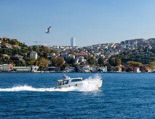 A sleek white boat, probably a private yacht, cuts through the crystal clear waters, leaving a trail of foam in its wake on Istanbul's Bosphorus. The iconic skyline stretches along the Asian side of t