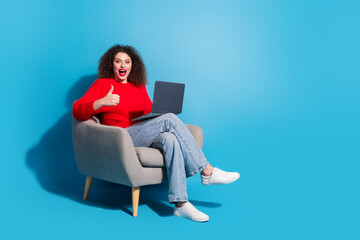 Full body photo of attractive young woman sit armchair hold netbook thumb up dressed stylish red clothes isolated on blue color background