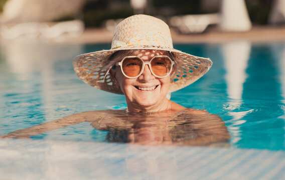 Portrait of smiling caucasian senior woman with hat and sunglasses relaxing into the outdoor swimming pool enjoying relax and vacation