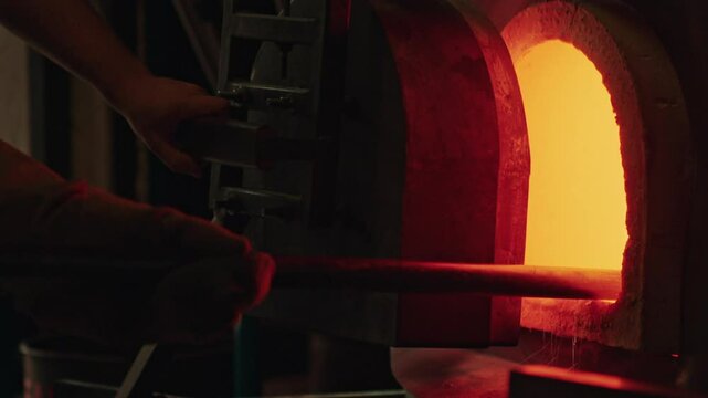 Shot of two workers pulling and trimming molten glass from a furnace in a glassblowing studio