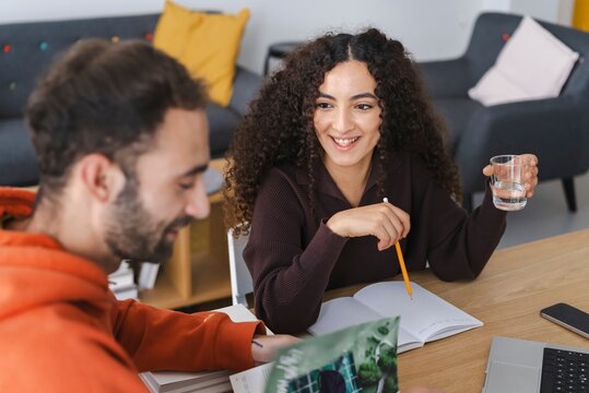 A woman with curly hair holds a pencil and glass of water, smiling and engaging in a conversation with a male colleague who is out of focus, sitting in a modern, comfortable office setting.