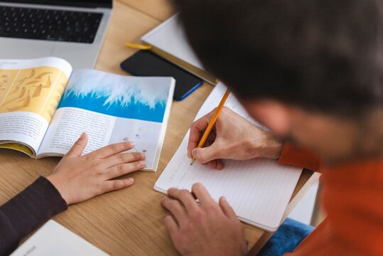Two people&rsquo;s hands are seen on an open book showing a close-up interaction as they discuss content in a relaxed, collaborative workspace with notebooks and a laptop nearby.