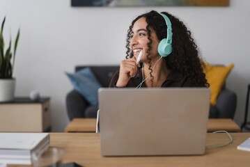 A smiling woman with curly hair and green headphones while working on a laptop in a modern workspace, giving a friendly, approachable vibe in a comfortable indoor environment.