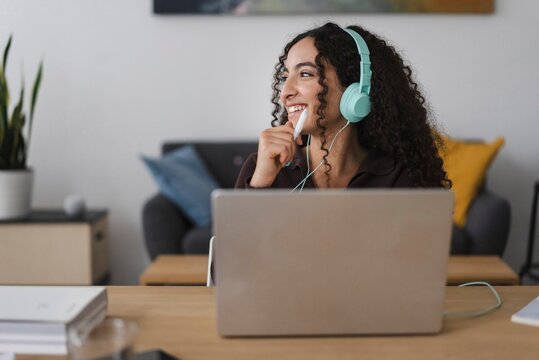 A smiling woman with curly hair and green headphones while working on a laptop in a modern workspace, giving a friendly, approachable vibe in a comfortable indoor environment.