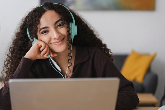 A smiling woman with curly hair and green headphones while working on a laptop in a modern workspace, giving a friendly, approachable vibe in a comfortable indoor environment.