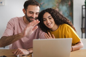 A woman in a yellow shirt and a man in a pink shirt smile as they video chat on a laptop together, appearing joyful and engaged in a relaxed indoor setting, creating a warm and friendly atmosphere.