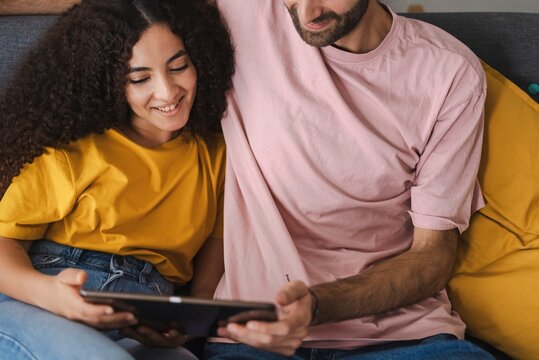 A couple sits close together on a sofa, smiling and looking at a tablet, sharing an intimate and relaxed moment in a cozy indoor setting that emphasizes warmth and connection.