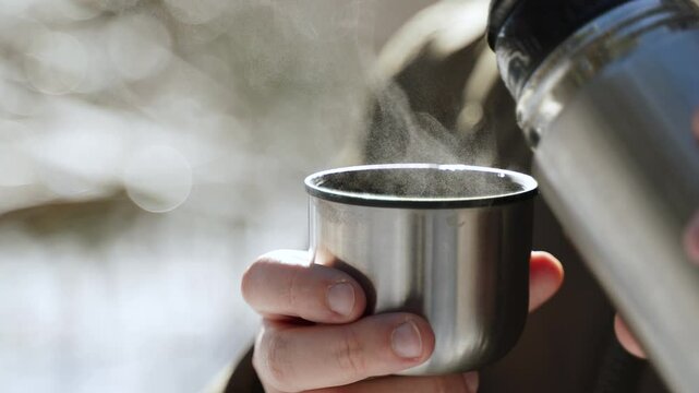 Close up of hand holding steaming stainless steel mug with hot beverage. Background is blurred, evoking warmth and comfort in cold setting. Female person on sunny winter day.