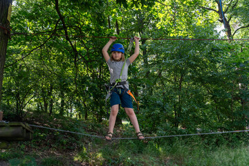 Young boy on rope bridge at adventure park with helmet and safety harness, balancing on ropes surrounded by lush green forest on sunny day.