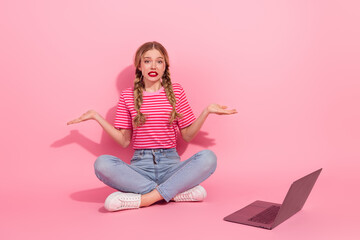 Confused young woman sitting cross-legged with braided hair and laptop on pink background