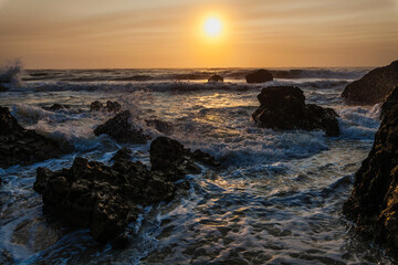 coastal sunset scene, waves rush in from the open sea, filling the space between the dark, rugged rocks