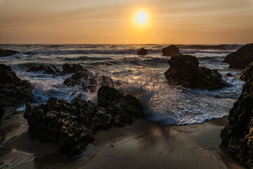coastal sunset scene, waves rush in from the open sea, filling the space between the dark, rugged rocks