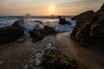 Obraz premium coastal sunset scene, waves rush in from the open sea, filling the space between the dark, rugged rocks