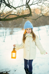 Girl in a blue hat holding a lantern against a snowy background. Magical winter atmosphere.